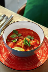 Spicy thai tom yum soup is served in blue bowl on red plate in restaurant