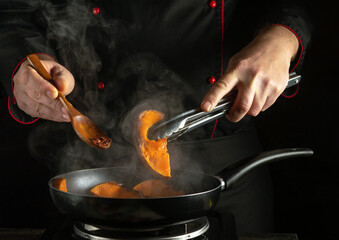A chef skillfully prepares sweet potato slices in a skillet, using tongs and a wooden spoon, as steam billows around in a warm kitchen environment during the evening