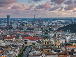 Obraz premium Aerial view of Budapest, Hungary, highlighting St. Stephen's Basilica, the Danube River, historic architecture, a skyscraper, and rolling hills.
