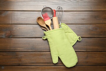 Different kitchen utensils in oven mitten on wooden table, top view