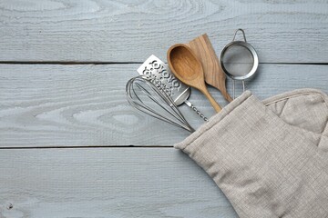 Different kitchen utensils in oven mitten on light grey wooden table, top view. Space for text