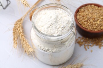Wheat flour in glass jar, grains and spikes on white table, above view