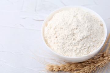 Wheat flour in bowl and spikes on white table, above view. Space for text