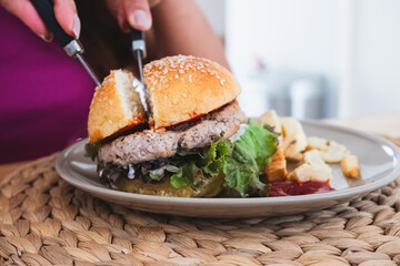 Adult woman's hands cutting a delicious homemade burger with lettuce and fries on a plate, ready to eat, comfort food.