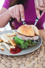 Adult woman cutting a plant-based burger with lettuce and a side of french fries and ketchup on a woven placemat, healthy meal.