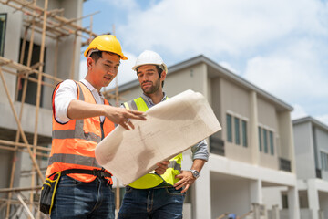 Asian male civil engineer and caucasian male architect wears safety vest with helmet discuss and look at blueprints in housing estate construction site