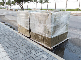 Stacked paving bricks wrapped in plastic on wooden pallets placed on a sidewalk by the roadside....