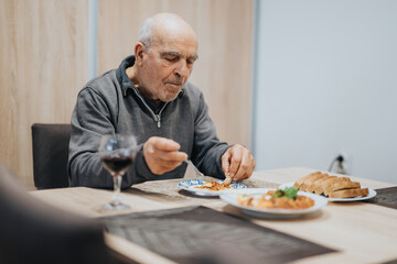 Elderly man seated at a dining table relishing a plate of food with bread on the side and a glass of red wine, representing warmth and tranquility.