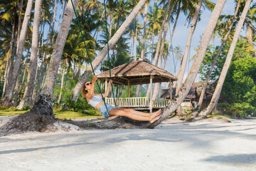Woman on swings at tropical beach, enjoying freedom and sunshine under palm trees, paradise vibes.