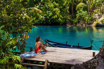 Woman in swimsuit sitting on a wooden pier by a turquoise lagoon, surrounded by lush tropical nature.