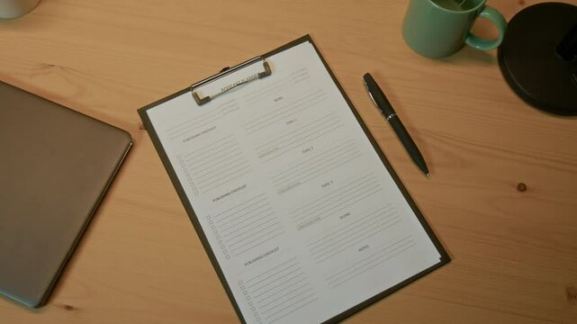 High angle shot of black clipboard with script of podcast, pen, laptop and pastel green cup on wooden table in studio