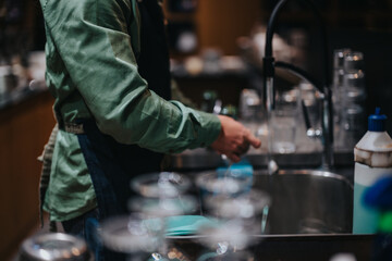 A bartender wearing an apron washes glasses at a sink in a cozy atmosphere.