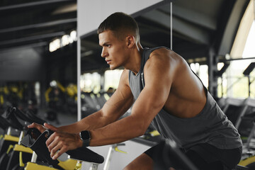 Fototapeta premium Side view of concentrated man cycling stationary bike in modern gym while holding handle bar tightly and looking forward. Fitness and health care concept.