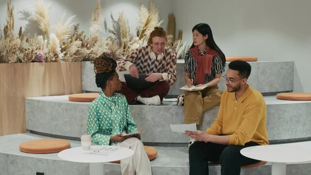 Full shot of diverse team of gen Z social media managers hanging out in cozy coworking workspace and discussing ideas for new mutual project, using documents and digital devices