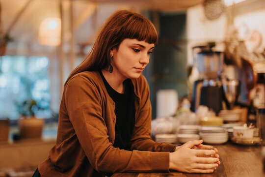 A thoughtful young woman sits alone in a warmly lit cafe, lost in deep contemplation. The mood is introspective and quiet, with a cozy and inviting atmosphere surrounding her.
