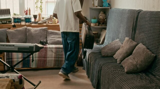 Back view of guy in stylish casual attire entering domestic room and moving towards couch with cushions, sitting down, opening case and taking out violin with fiddlestick