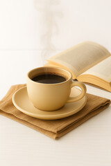 A steaming cup of black coffee in a yellow ceramic mug on a saucer. A brown napkin and an open book are placed beside it on a light wooden surface.