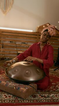 Vertical shot of young African American woman sitting on carpet on the floor of home studio and beating metallic glucophone with drumsticks while creating new ethnic native music