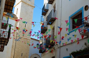 Vibrant pennants in bright colors sway gently above a narrow street lined with whitewashed buildings and wrought iron balconies