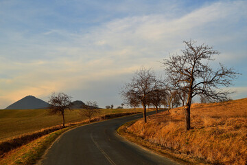 Fototapeta premium Sunlit Country Road Curving Through Hills Beneath Evening Clouds