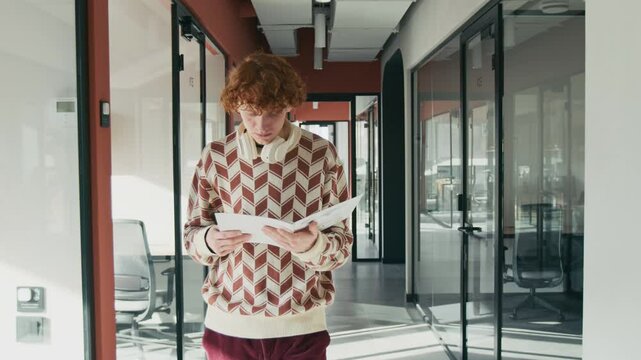 Tracking shot of young adult Caucasian man with ginger hair walking down corridor in coworking space, reading documents and greeting female colleague, passing by offices with glass walls