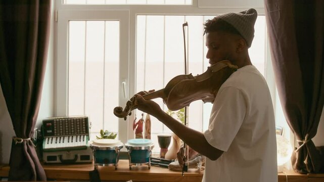 Push in of young male musician in casual apparel standing in modern home studio against window and playing classical music on violin during repetition