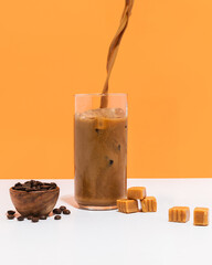 Caramel coffee being poured into glass of ice with coffee and caramels around on orange and white studio background