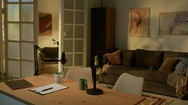 Slow motion of spacious studio illumed by warm sunrays. Wooden table with set professional microphones, laptop, two cups and script on top
