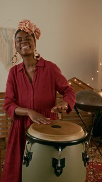 Vertical shot of cheerful female musician in ethnic accessories and casual dress beating percussion energetically while creating new music and enjoying repetition