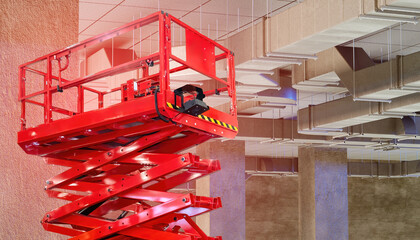 Red scissor lift platform positioned under ceiling ductwork in an unfinished building interior, used for HVAC or maintenance work. Activity: Construction, HVAC installation.