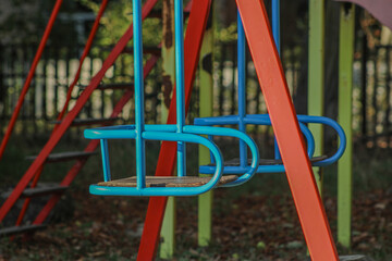 A playground swing set with colorful metal frames, including blue seats and red supports. The vibrant hues and simple design suggest a welcoming play area