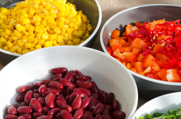 Close-up of several white and grey bowls filled with fresh, colorfully sliced ​​green onions, red beans, diced red peppers, yellow sweet corn and green peas ready for cooking.