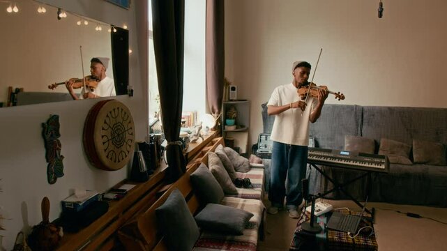 Full length shot of young musician in jeans and t-shirt playing violin while standing by couches with cushions in home studio and preparing for concert of classical music