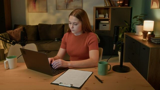 Teen girl looking at script of interview on paper and typing on keyboard to cope text, she sitting at desk surrounded by professional microphones