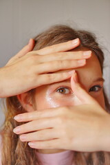 Charming young girl playfully covering her face with hands, showcasing a radiant glow and her expressive eye