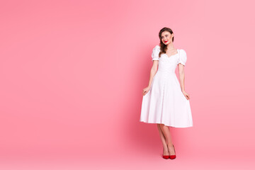 Young woman in an elegant white dress and red shoes posing against a soft pink background with charming confidence