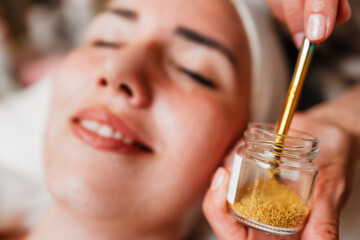Beautician carefully applying gold particles with a brush on the forehead of a woman lying with closed eyes, during a rejuvenating treatment in a beauty spa, enhancing skincare and relaxation