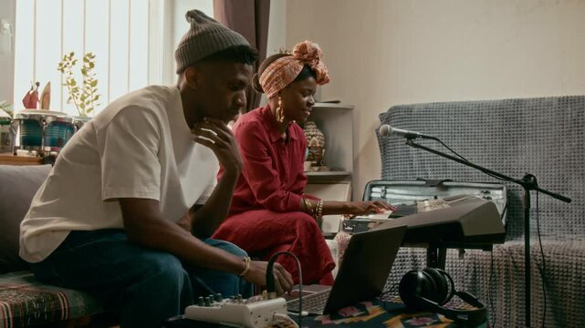 Side view of two young African American musicians in casual attire sitting on sofa in home studio while one of them playing synthesizer piano and consulting with guy recording music