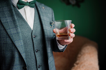 Stylish man in a green tweed suit with a bowtie holds a glass of whiskey, enjoying a drink in an elegant setting.