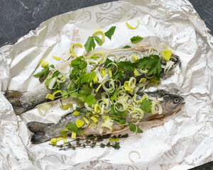 Fresh whole fish with leeks and herbs wrapped in foil on dark surface