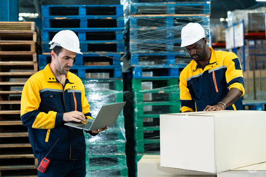 Warehouse teamwork between african adult male and caucasian adult male workers wearing uniform and helmet during packing and checking parcels using laptop for shipment preparation process