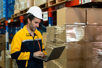 warehouse employee updating inventory record on laptop while standing near stacked cardboard boxes in aisle wearing safety uniform and white helmet inside stockroom