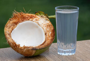 Fresh green coconut water in glass on wooden table, nature background. Half-cut coconut with juicy...