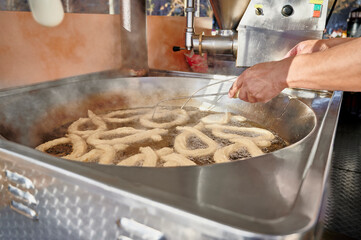Hands of a churro vendor flipping porras with metal rods in boiling oil inside an industrial deep fryer. Traditional skills and tools used in Spanish churro-making