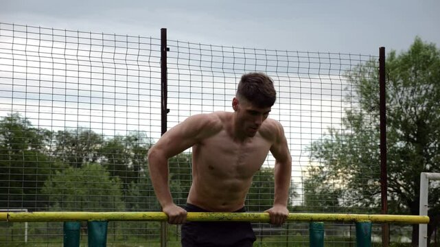 Male shirtless athlete doing push ups on parallel bars at nature. Strong sporty man exercising at sports ground. Young sportsman training hard outdoor. Concept of active sportive lifestyle. Slow mo