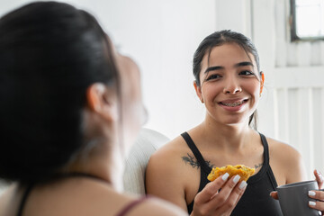 young latina woman laughing while looking at her friend, eating her snack, woman with braces,...