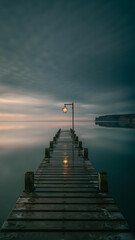 Obraz premium Wooden pier stretching into the calm sea under a cloudy sky at dusk with a lamp post