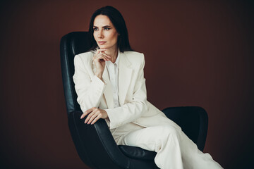 Elegant businesswoman in a white suit sitting confidently in a chair with a thoughtful gesture in a...