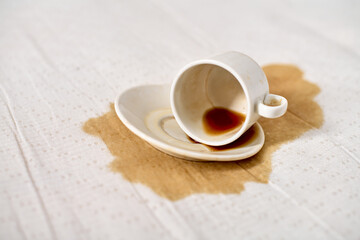 Empty coffee cup tipped on white tablecloth, creating brown liquid stain. Soft lighting accentuates stark contrast between white fabric and dark spill, conveying sense of accident or clumsiness