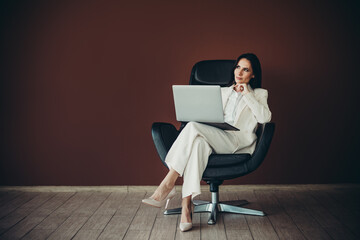 Stunning businesswoman sitting in office chair with laptop against brown background, portraying...